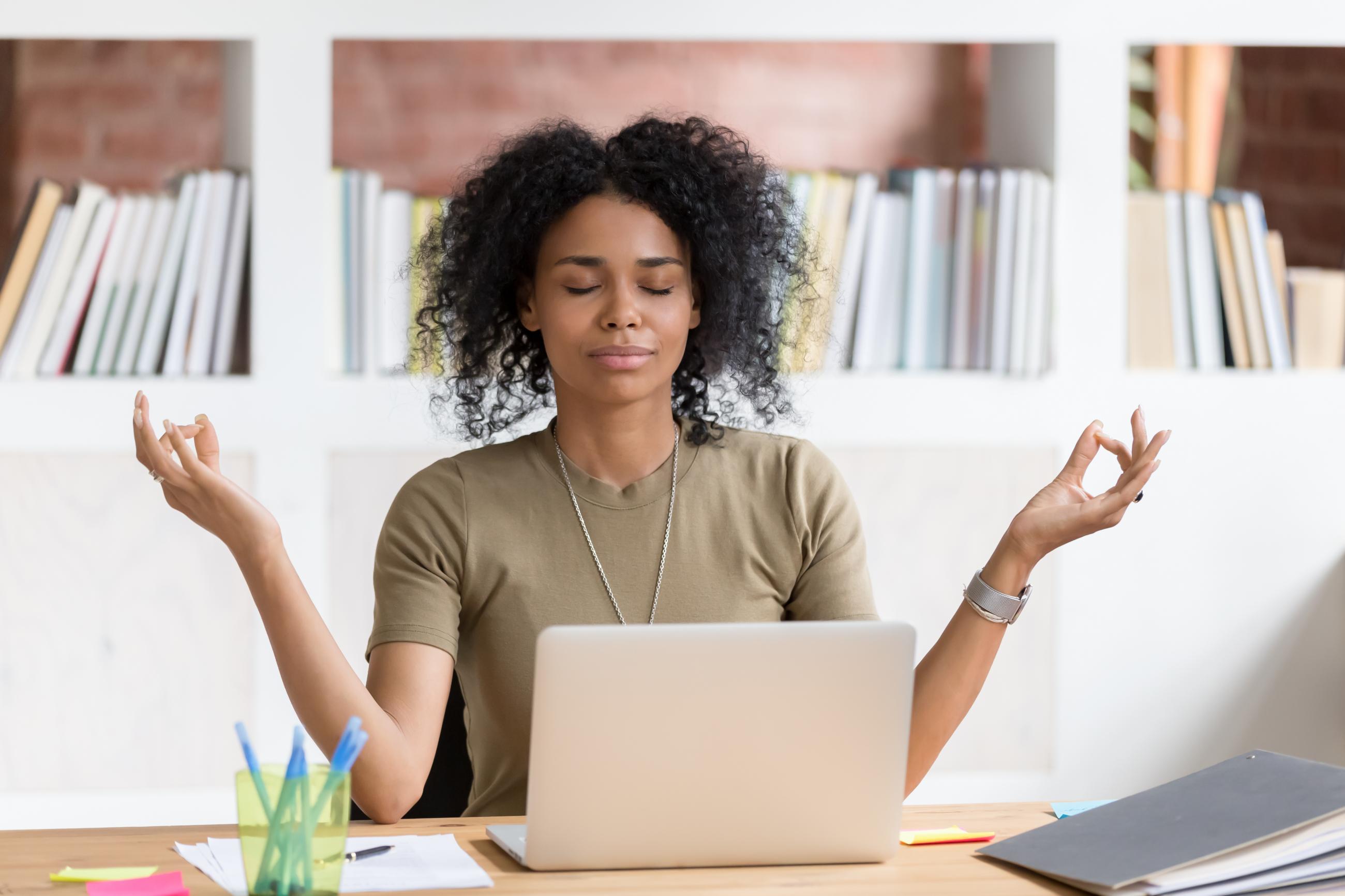 An African-American woman is meditating while sitting at work desk.